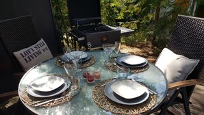 A round glass table set for four with plates, cutlery, and glasses, surrounded by wicker chairs and a grill in the background. - Naturally happy (170m to the Müritz) (Röbel/Müritz)