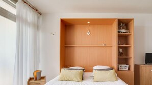 Une chambre à coucher comprenant un lit, deux oreillers, une table de chevet, une commode, une étagère avec des objets décoratifs et un téléviseur posé sur une armoire en bois.