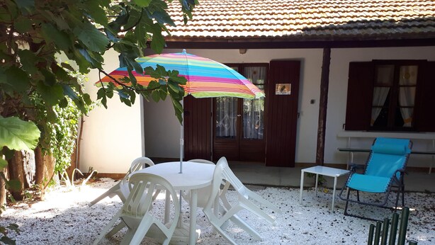 Une terrasse avec une table ronde et des chaises, un parasol coloré, un fauteuil bleu et un mur blanc surmonté d’un toit en tuiles.