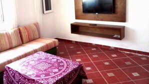 Ein Zimmer mit an der Holzwand befestigtem Fernseher, einer gestreiften Kissenbank und einem kleinen Tisch mit einem Tischläufer mit Muster. Der Boden ist mit roten, gemusterten Fliesen belegt.