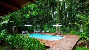 A wooden deck surrounding a swimming pool with white lounge chairs and umbrellas, set in a lush, green garden. - Casa Cerise (Cahuita)