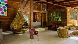 A cozy cabin interior with a large window, a woven hanging chair, a cushioned bench, and a colorful wall art piece. - Casa Cerise (Cahuita)
