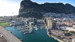 Un paisaje urbano costero con un puerto deportivo lleno de barcos, rodeado de edificios residenciales y comerciales, y un acantilado rocoso destacado al fondo.