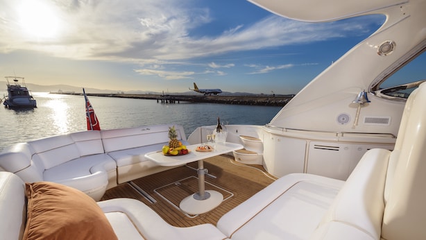 Interior de un yate con un sofá de cuero blanco, una mesita con una botella y frutas, y una vista de un muelle y un avión al fondo.