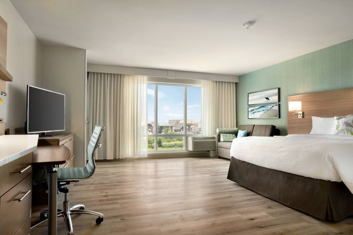A hotel room with a large bed, a desk with a computer, a chair, a window with curtains, and a view of buildings outside.