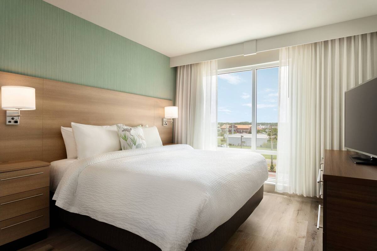 A hotel room with a large bed, a TV, a wooden nightstand, and a view of the city through the window.