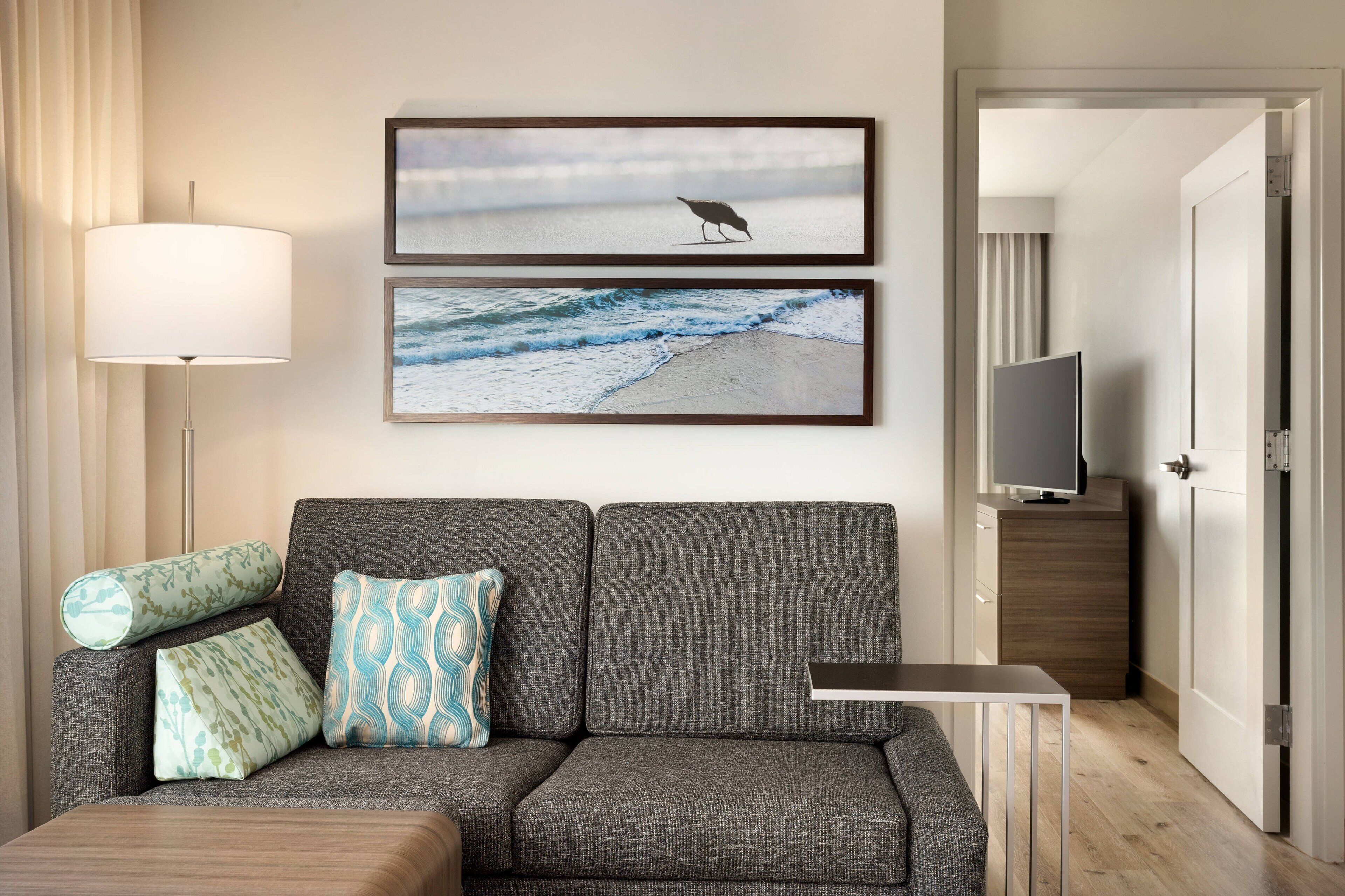 A modern living room with a grey sofa, two patterned cushions, a side table, a flat-screen TV, and two framed pictures of a bird and a beach scene.