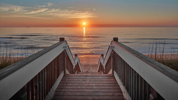Une promenade en bois menant à une plage, avec un coucher de soleil en arrière-plan.