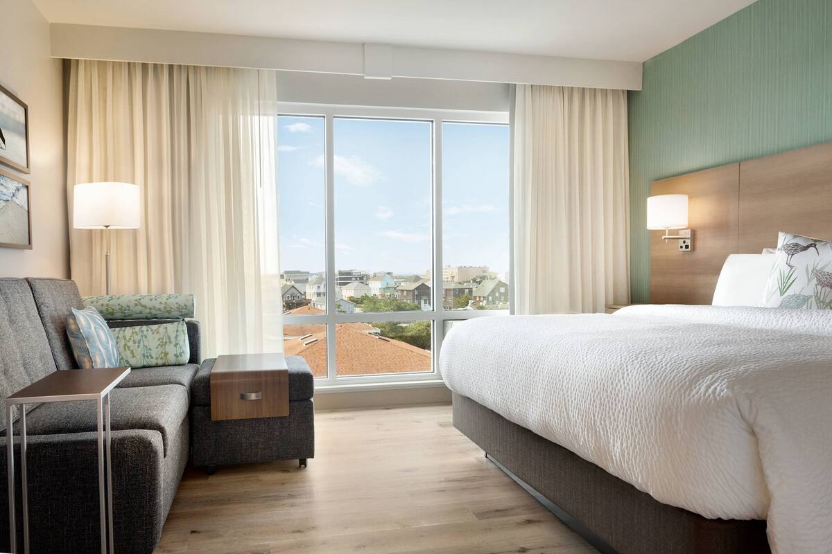 A hotel room with a large bed, a sofa, a small table, and a view of the city through large windows.