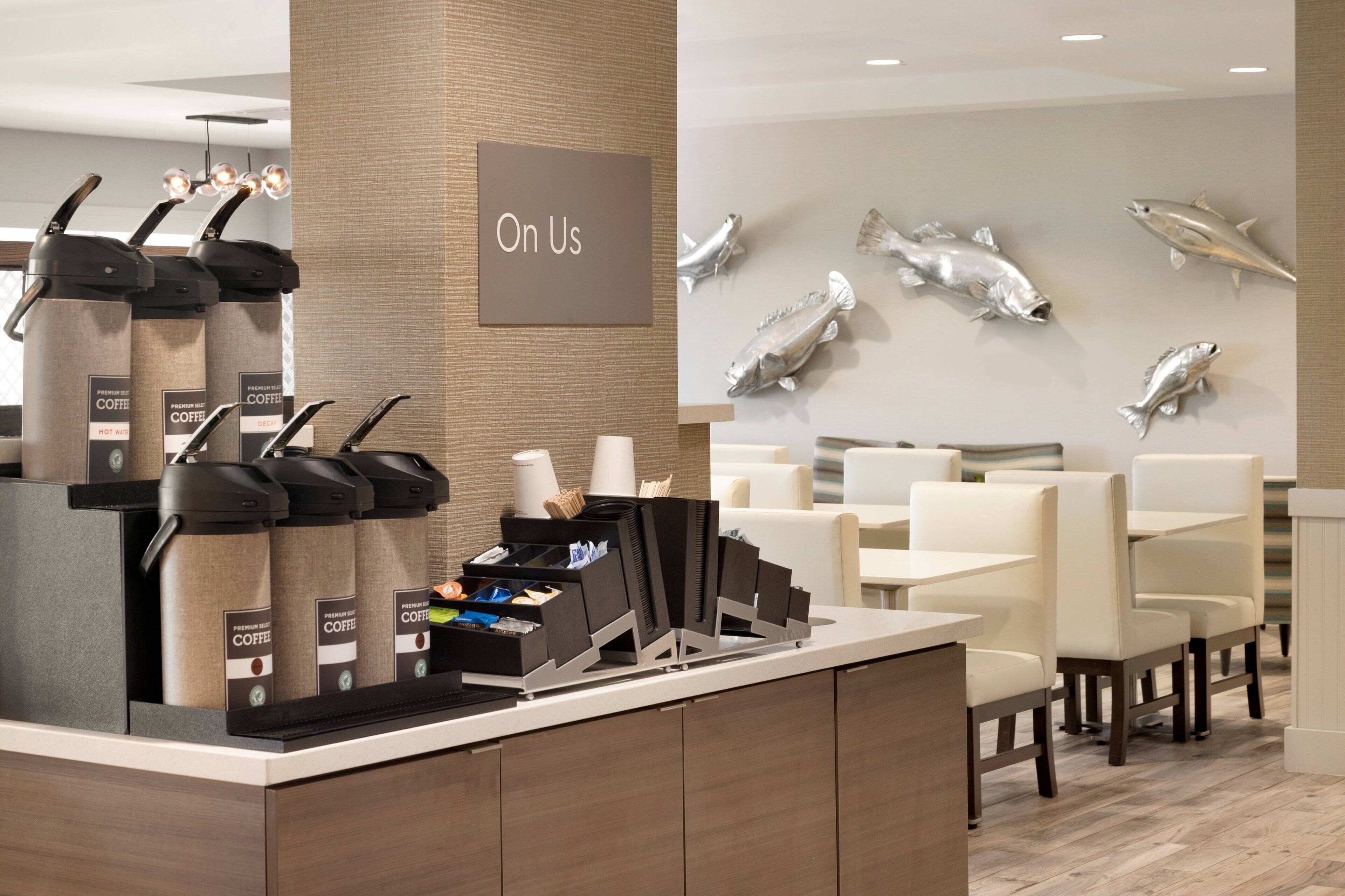Un espace café moderne doté de plusieurs distributeurs, d’une pancarte indiquant « Nous vous offrons » et d’un mur décoré de sculptures de poissons.