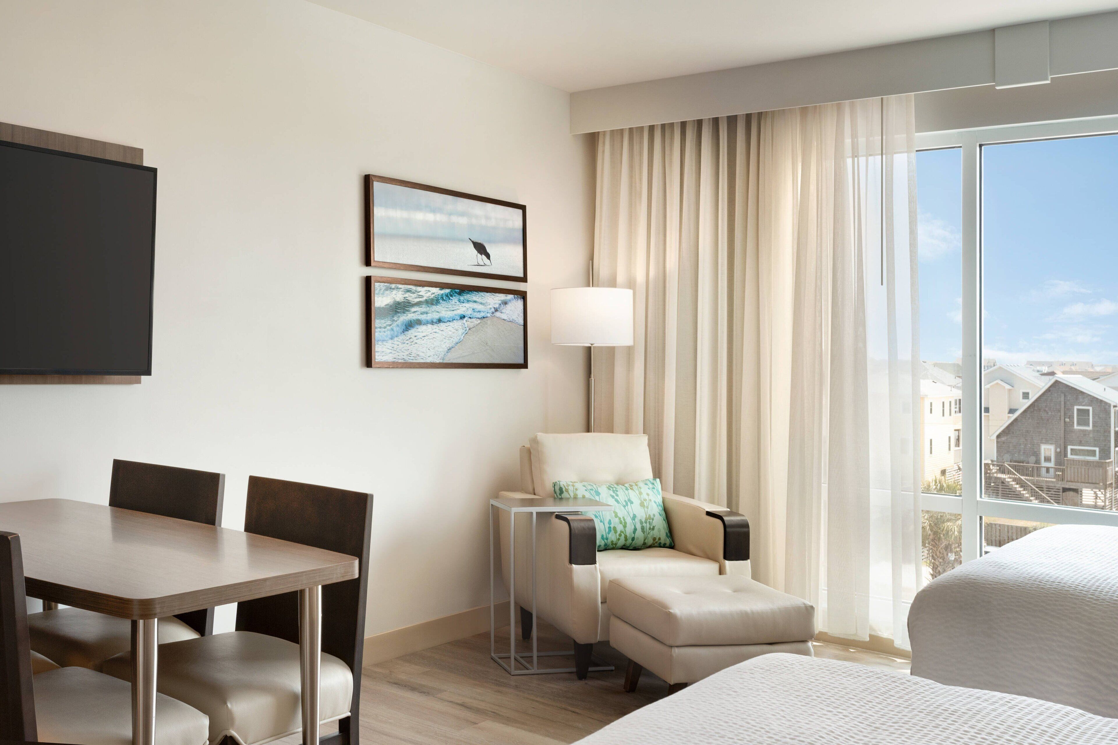 A hotel room with a dining table, two chairs, a sofa, a lamp, and a large window with a view of buildings.