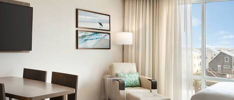 A hotel room with a dining table, two chairs, a sofa, a lamp, and a large window with a view of buildings.
