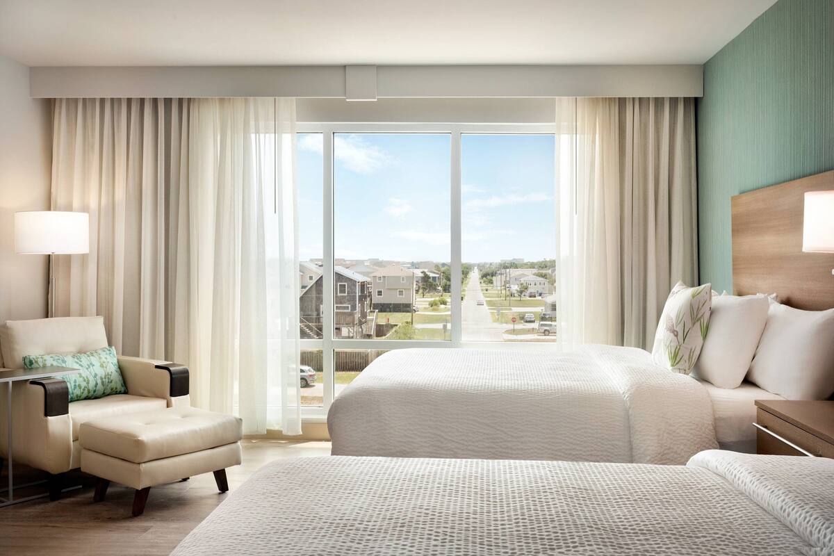 A hotel room with a large bed, a chair, a desk, and a view of a residential area through large windows.