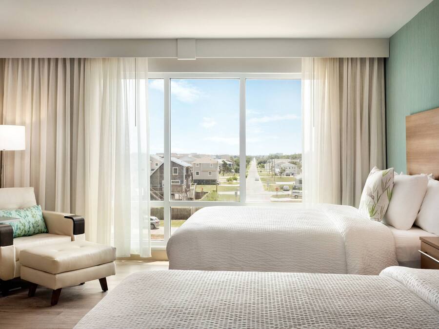 A hotel room with a large bed, a chair, a desk, and a view of a residential area through large windows.