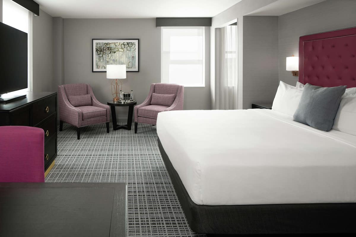 A hotel room with a large bed, two armchairs, a small table with a lamp, a TV, and artwork on the wall.