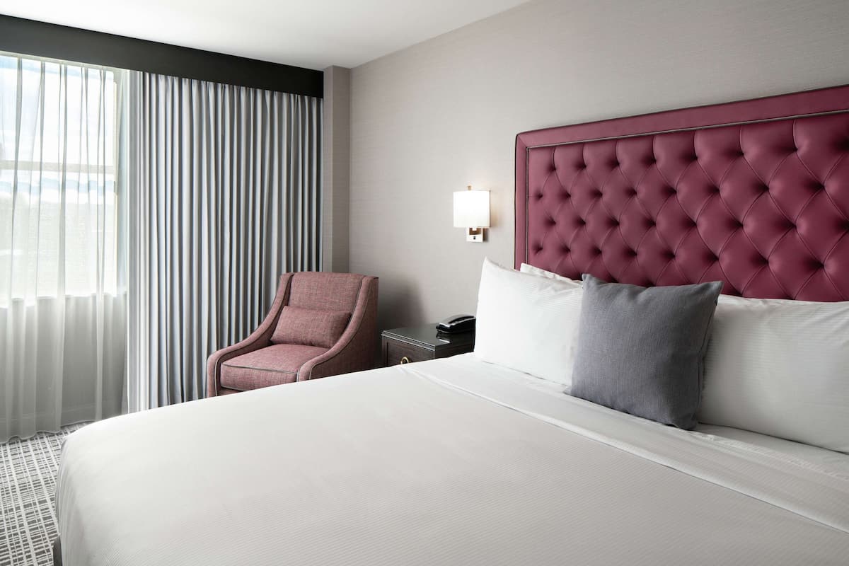 A hotel room with a large bed, a tufted headboard, a pink armchair, a bedside table with a lamp, and a view of the city through sheer curtains.