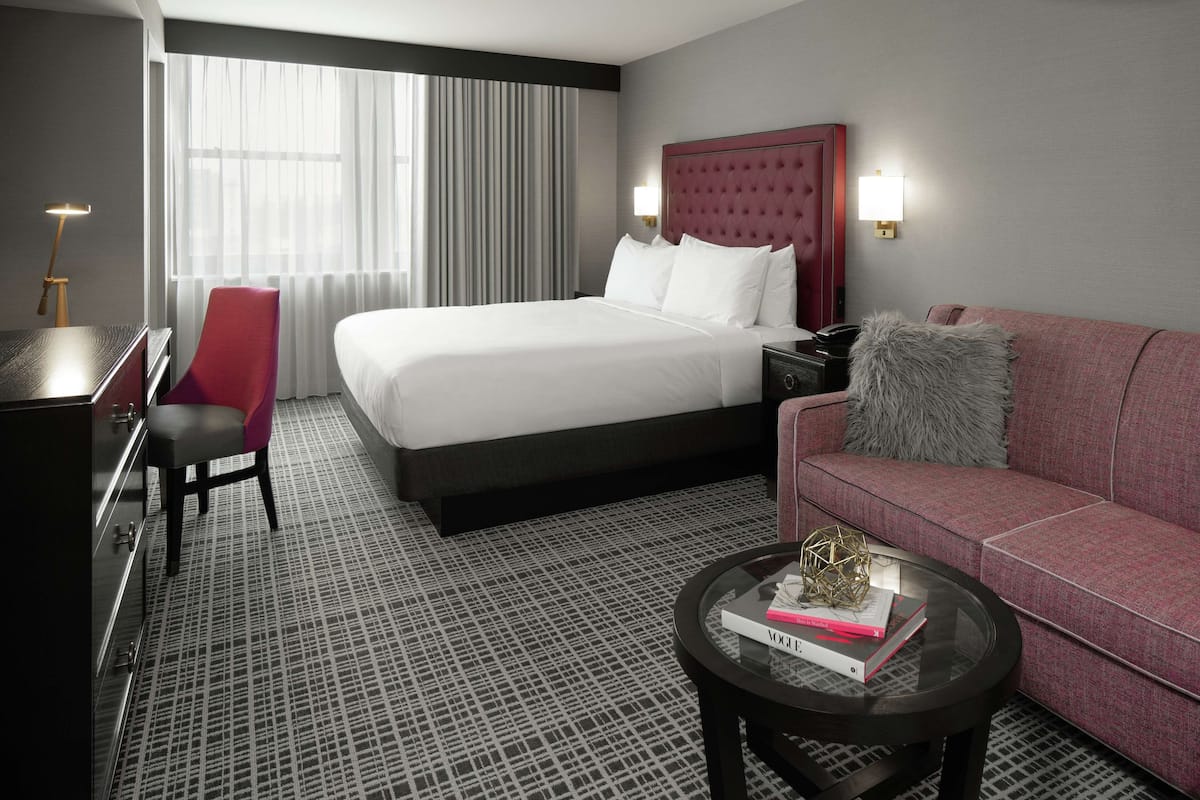 A hotel room with a large bed, a red armchair, a sofa, a glass coffee table, and a desk with a lamp.