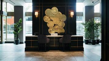 A lobby with a reception desk, two chairs, a wall-mounted art piece, and potted plants.