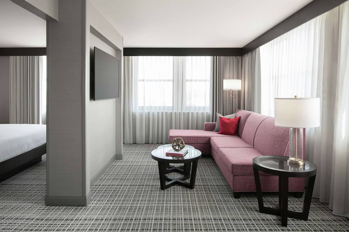 A hotel room with a bed, a pink sofa, two round tables, a TV, and a large window with curtains.