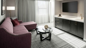 A modern hotel room with a grey sofa, a round coffee table, a flat-screen TV, and a large window with curtains. - The Axis Moline Hotel, Tapestry Collection by Hilton (Moline)