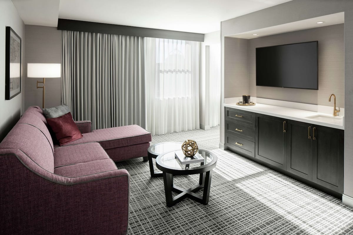 A modern hotel room with a grey sofa, a round coffee table, a flat-screen TV, and a large window with curtains.