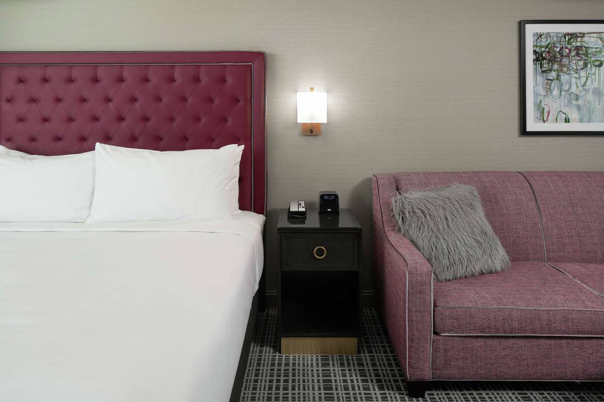 A hotel room with a tufted headboard, a bedside table with a lamp, a clock, and a sofa with a fluffy pillow.