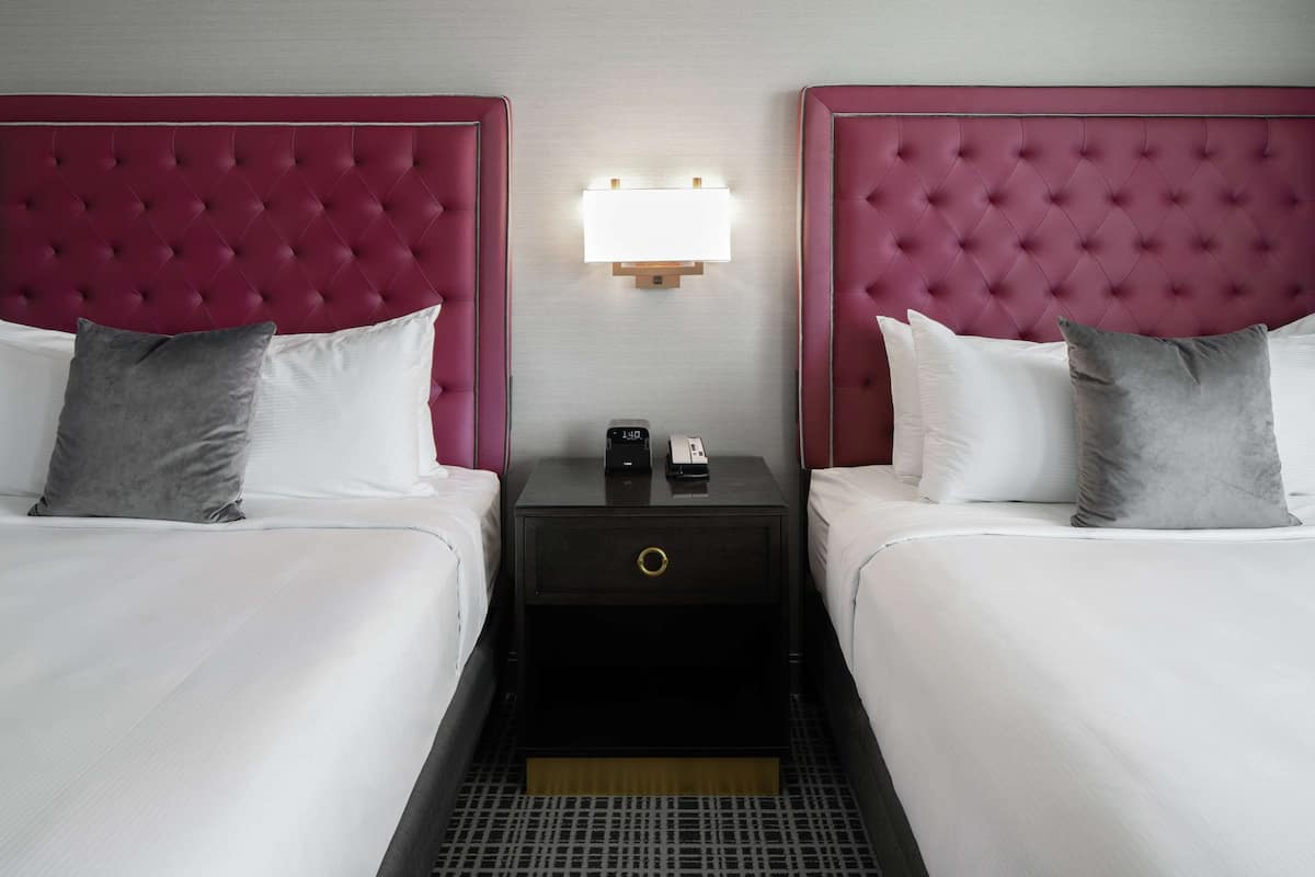 A hotel room with two beds, each with a tufted headboard, a bedside table with a lamp, and a small drawer.