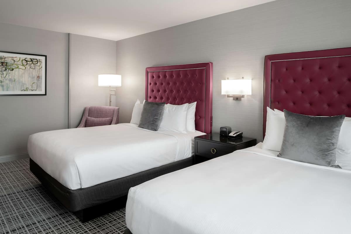 A hotel room with two beds, each with a tufted headboard, white bedding, and a nightstand with a lamp.