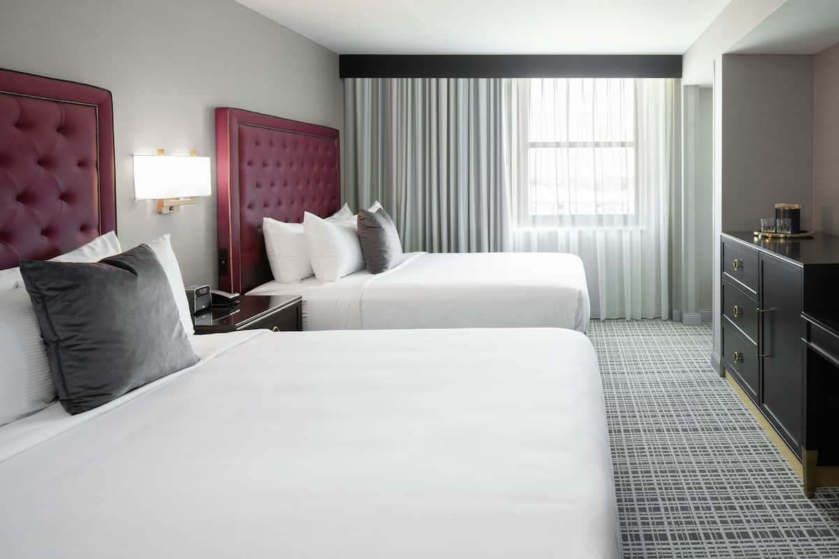 A hotel room with two beds, a nightstand, a dresser, and a large window with curtains.