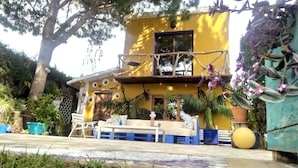A yellow building with a balcony, surrounded by various plants and a seating area with a table and chairs. - Comfortable apartment and swimming pool in the shade of the pine forest of Chipiona (Chipiona)
