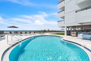 Piscina com espreguiçadeiras e guarda-sóis, rodeada por um edifício moderno com varandas.