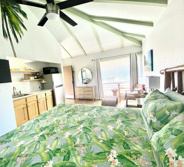 Une chambre avec un lit, un ventilateur de plafond, un coin cuisine et une fenêtre avec des rideaux.
