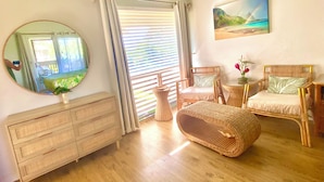 Una habitación con piso de madera, un aparador de madera, sillas de mimbre, un reposapiés de mimbre, un espejo y un cuadro de un paisaje.