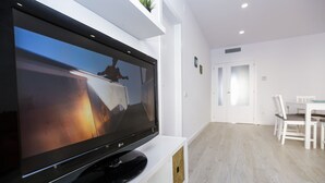 Een flatscreen-tv op een witte standaard in een moderne woonkamer, met een eethoek op de achtergrond.