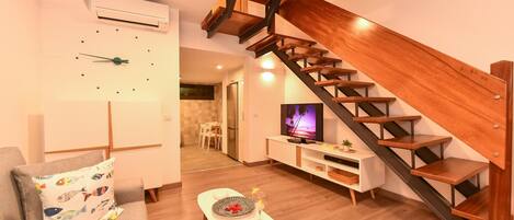 A modern living room with a wooden staircase, a flat-screen TV, and a dining area.