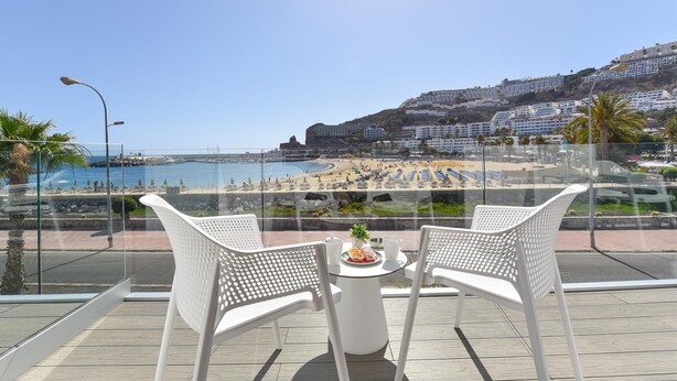 A balcony with two white chairs and a small table, overlooking a beach and a cityscape. - Morea Suites 3 by VillaGranCanaria (Puerto Rico)