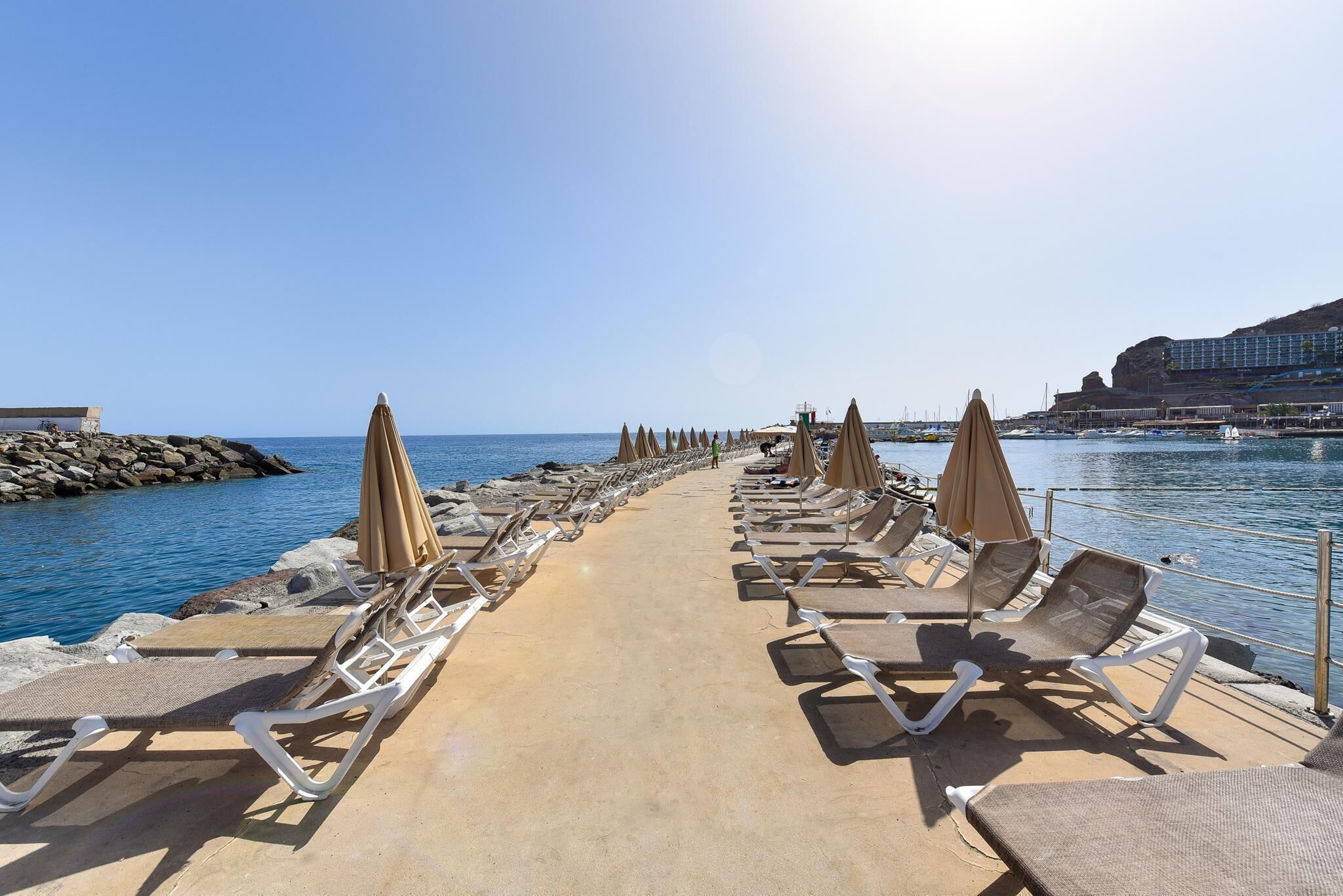 A sunlit promenade with rows of lounge chairs and umbrellas along a waterfront, leading to a rocky shoreline.