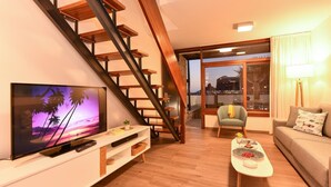 A modern living room with a wooden staircase, a flat-screen TV, a sofa, a coffee table, and a dining area with a lamp. - Morea Suites 3 by VillaGranCanaria (Puerto Rico)