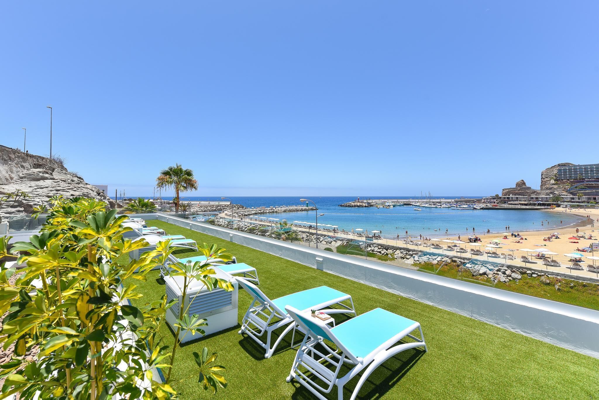 Un hôtel en bord de mer doté d’une terrasse verdoyante, de chaises longues et offrant une vue sur la plage et la mer.