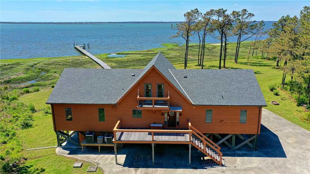 A two-story wooden house with a dock extending into a lake, surrounded by trees and grassy areas.