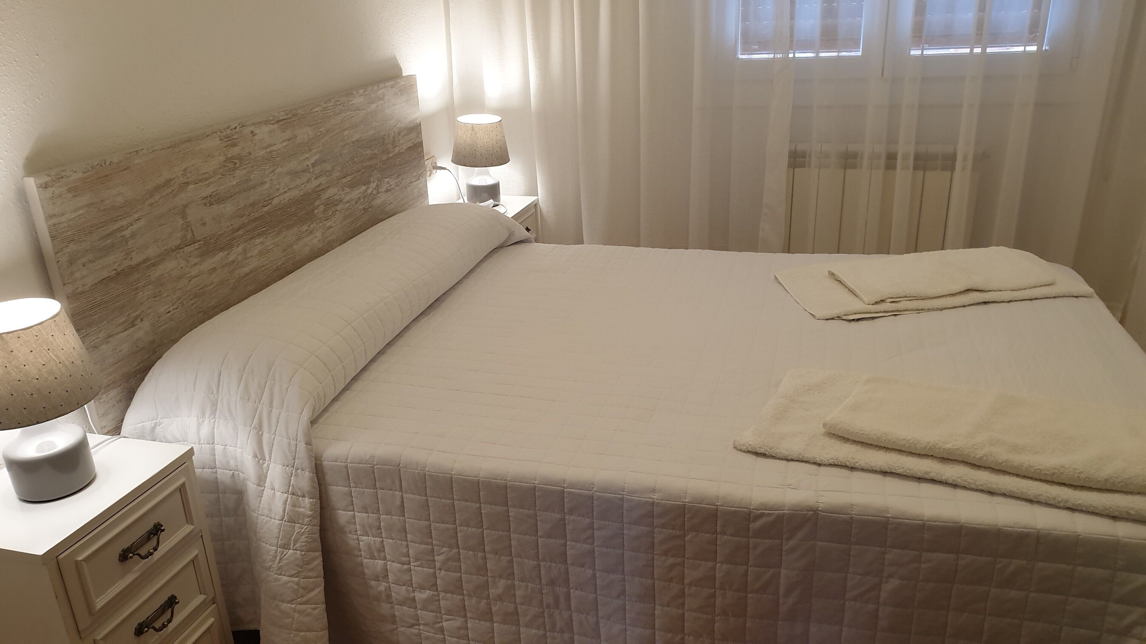 A neatly made bed with a quilted white duvet, a bedside lamp, and a white nightstand with drawers.