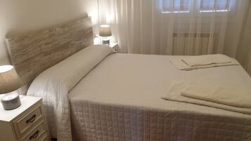 A neatly made bed with a quilted white duvet, a bedside lamp, and a white nightstand with drawers.