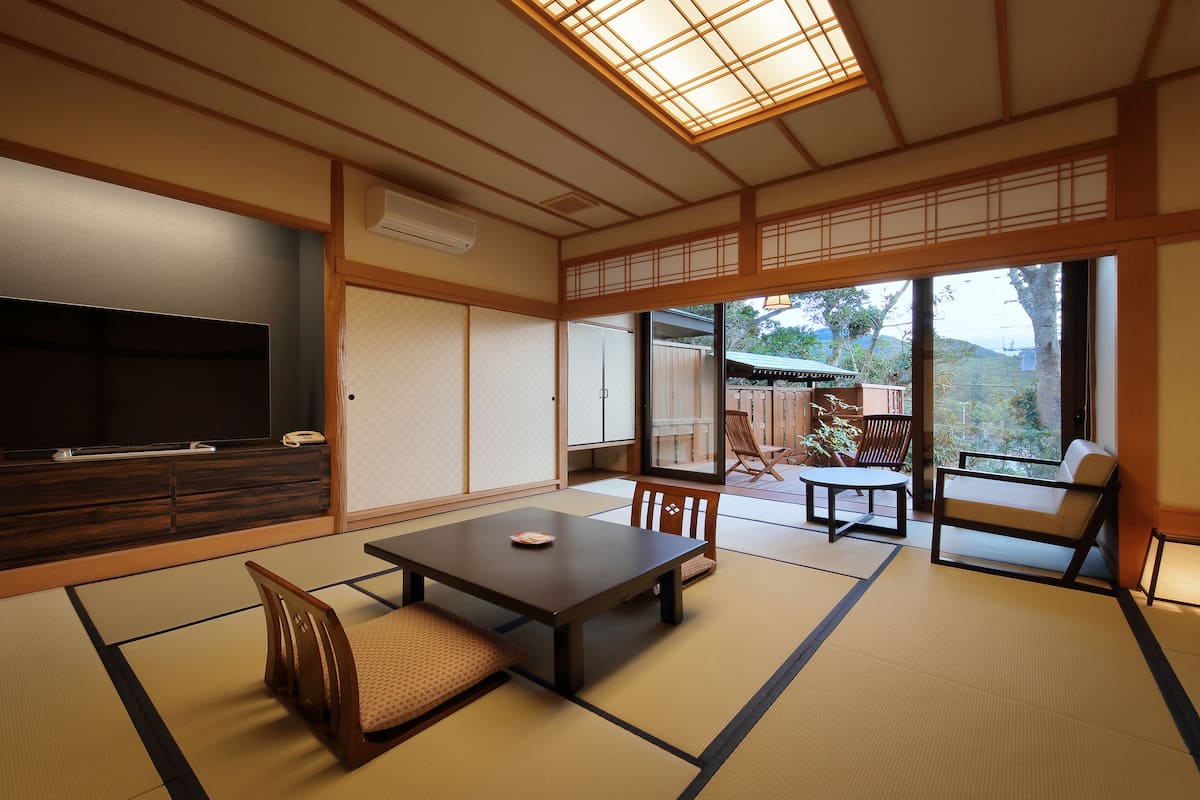 A traditional Japanese room with tatami flooring, a low table, chairs, a TV, and a sliding door leading to an outdoor area.