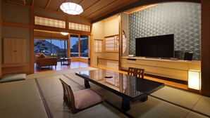 Una habitación tradicional japonesa con suelo de tatami, una mesa baja con cojines, un televisor y una puerta corredera que da a un balcón con vistas a las montañas.