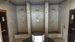 Un baño con tres cabinas de ducha, cada una con un cabezal y un panel de control. El suelo está cubierto de azulejos, y hay una zona de lavamanos central con encimera negra.