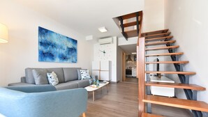 Ein modernes Wohnzimmer mit einem grauen Sofa, einem blauen Sessel und einem Couchtisch. Eine Holztreppe führt zu einer oberen Ebene.