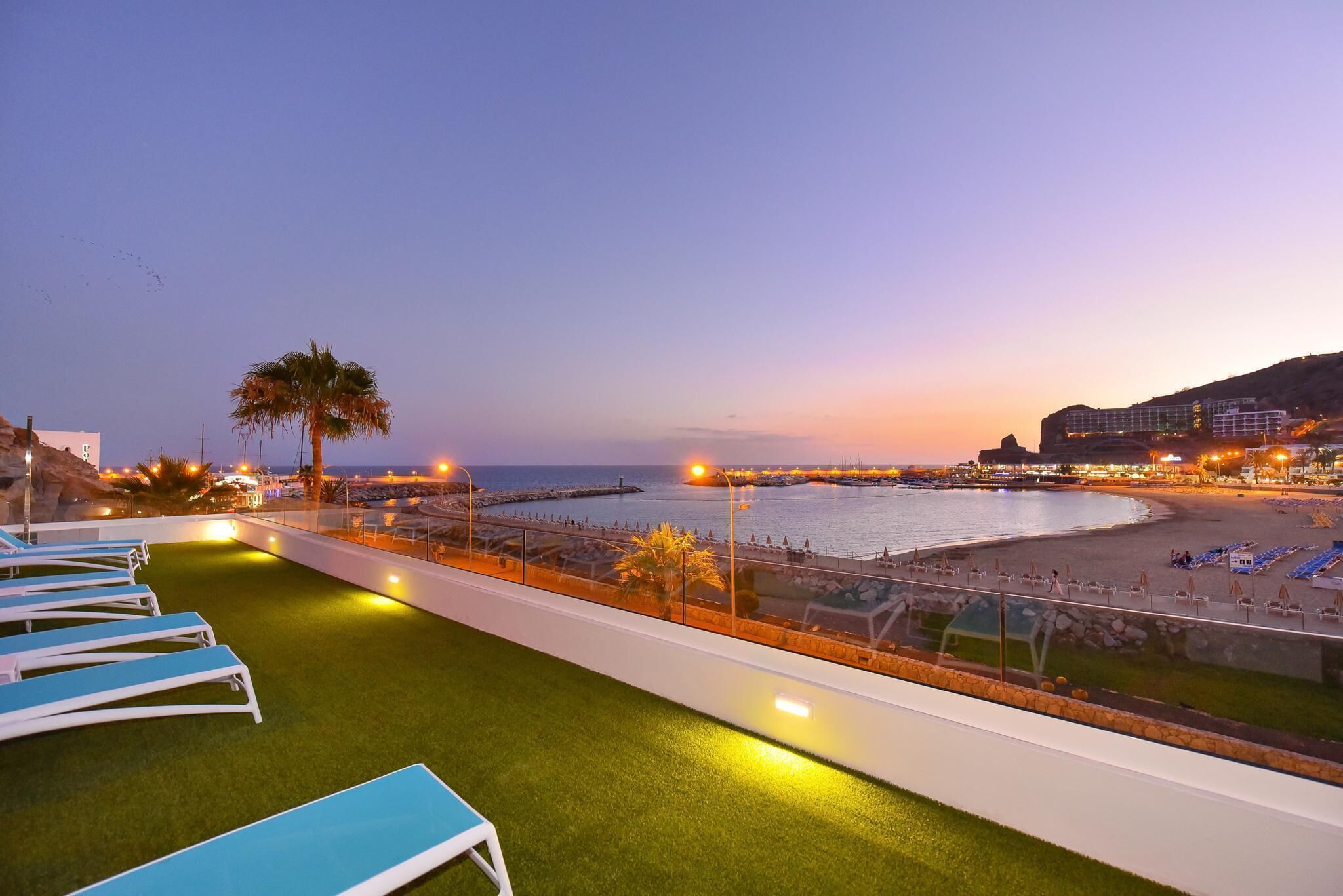 Een terras op het dak met kunstgras en lounge stoelen, met uitzicht op een strand en een pier in de zonsondergang.