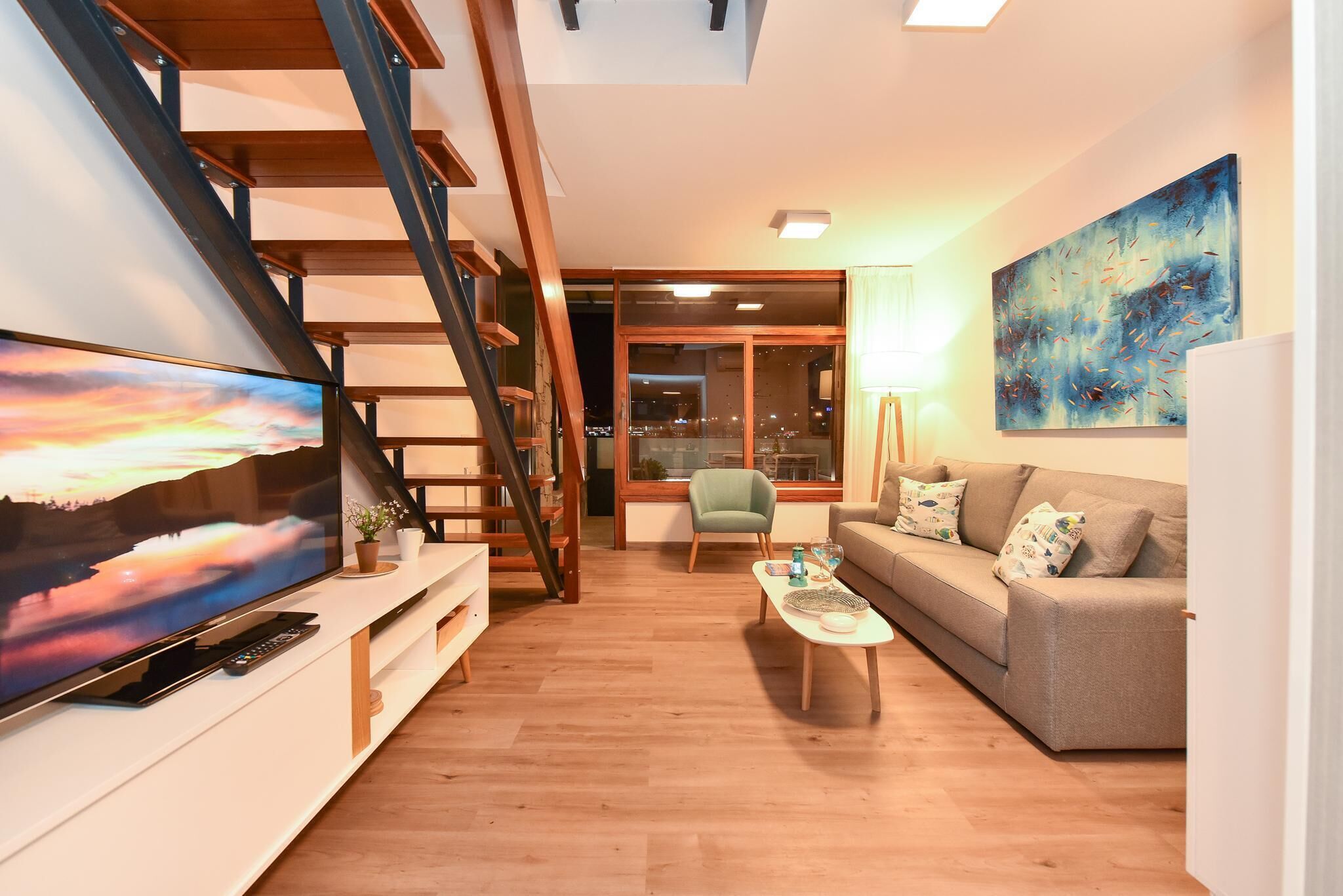 Una sala de estar moderna con un televisor de pantalla plana grande, una escalera, un sofá, una mesa de centro y un cuadro en la pared.
