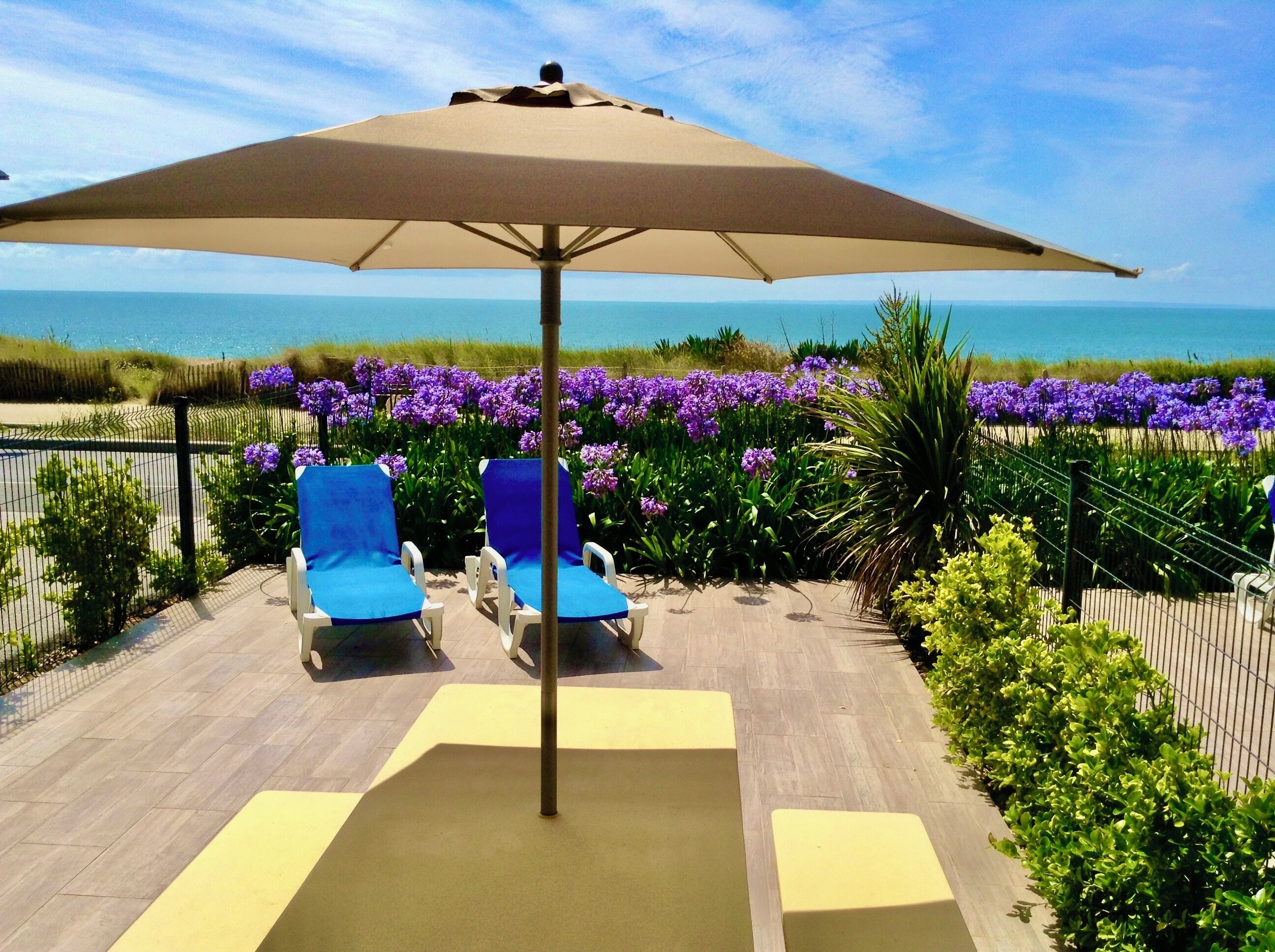 Une terrasse avec deux fauteuils de jardin bleus, un parasol beige et une vue sur l’océan.