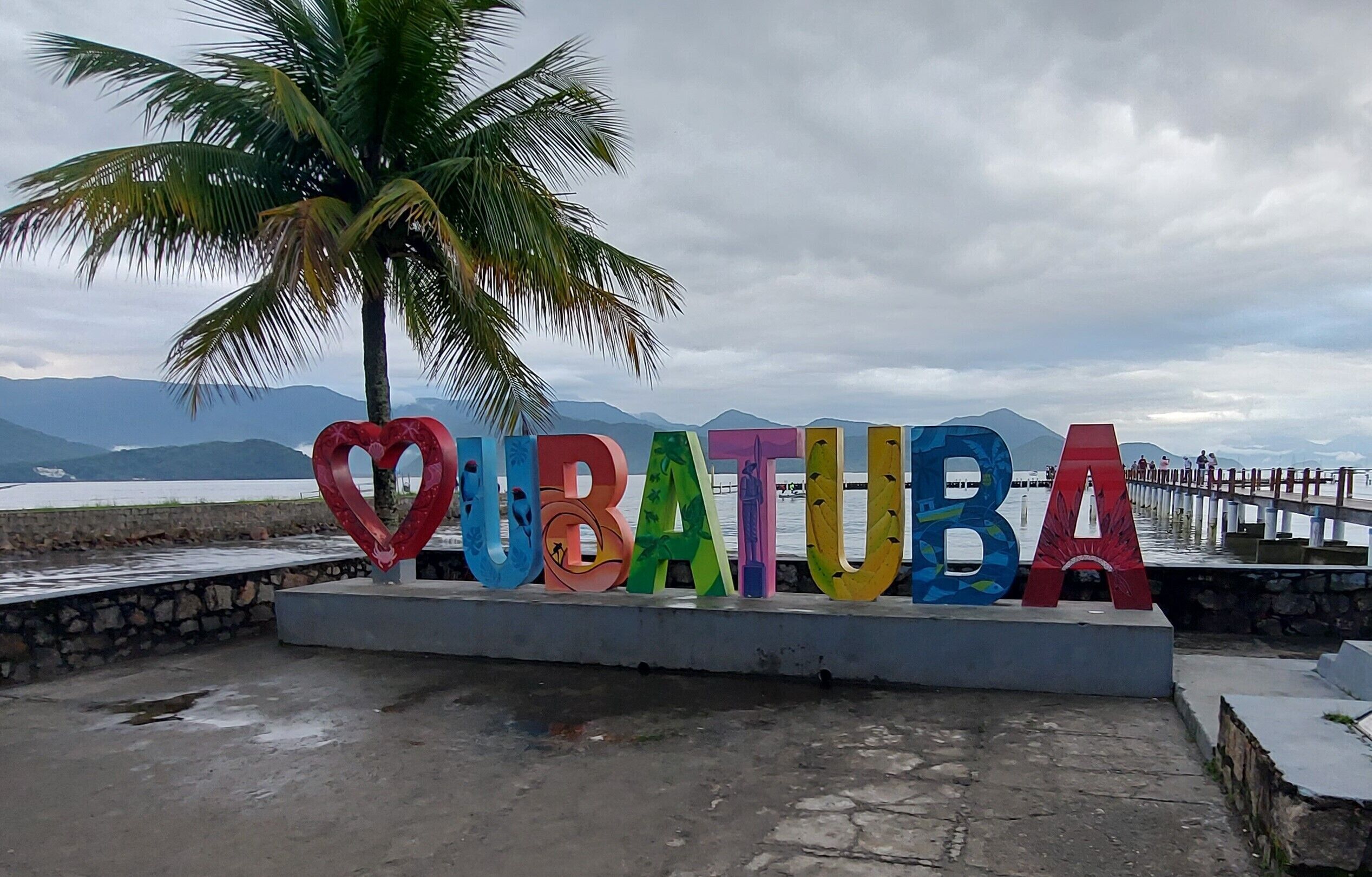 A sign with the word "QUBATUBA" in colorful letters, a palm tree, and a view of mountains and water.